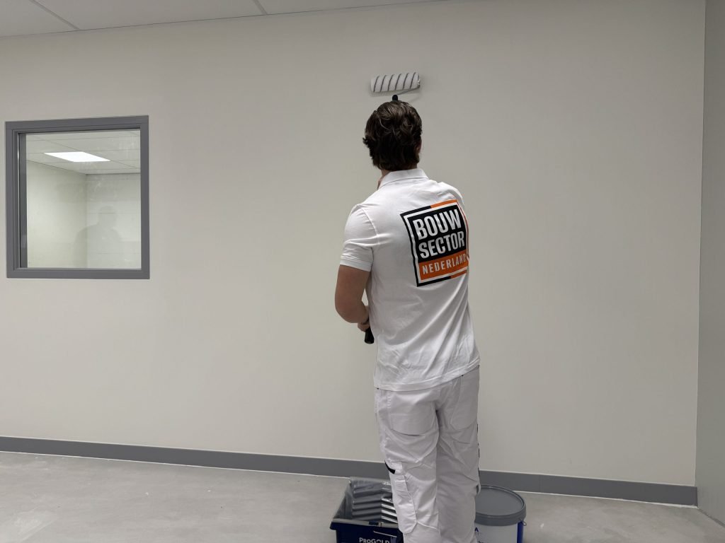 Schilder in witte werkkleding saust een lichte muur, met verfbak en emmer op de vloer