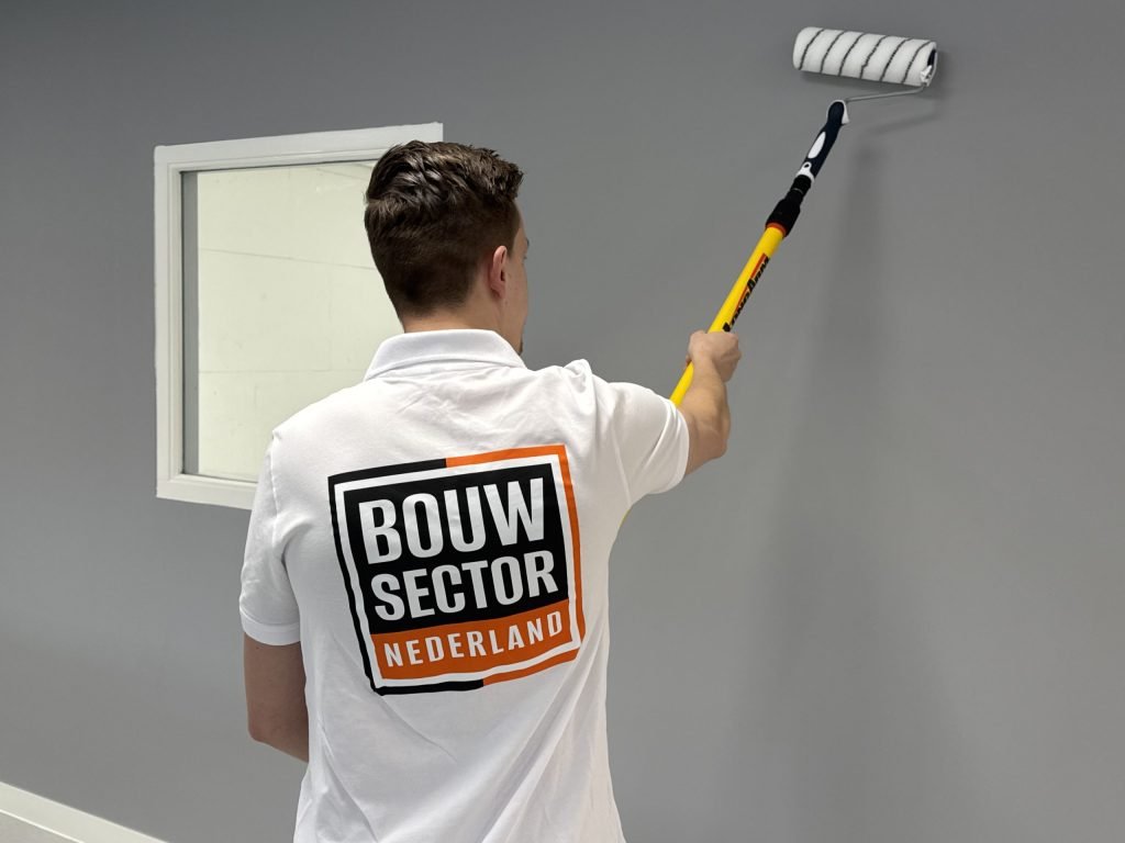 Schilder van Bouwsector Nederland zet een grijze wand in de verf met een roller naast een binnenraam