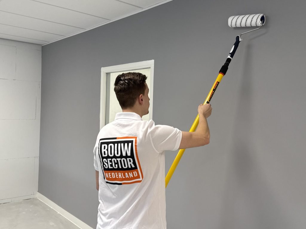 Schilder met Bouwsector Nederland-logo op zijn rug rolt een grijze wand naast een binnenraam