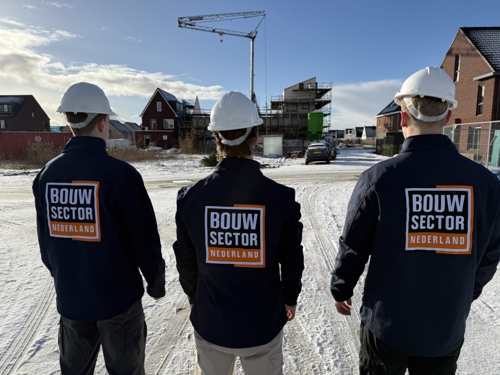 Drie vakmensen van Bouwsector Nederland met witte bouwhelm en jas met logo kijken over een besneeuwde straat naar bakstenen huizen