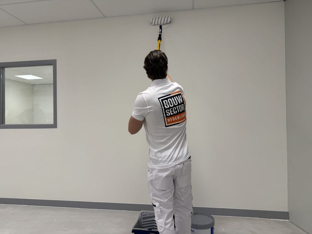 Schilder staat tussen verfbak en emmer en rolt met een verlengsteel een witte muur tot vlak onder het plafond