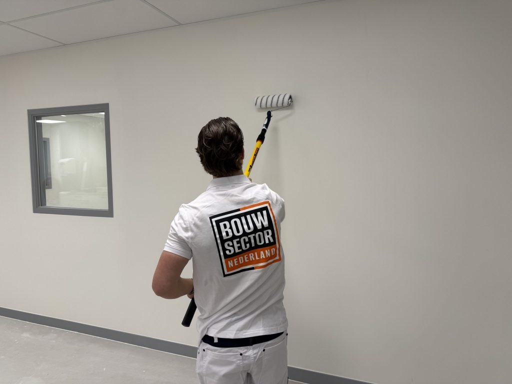 Schilder met het Bouwsector-logo op zijn rug rolt een lichte wand in een lege ruimte met een binnenraam